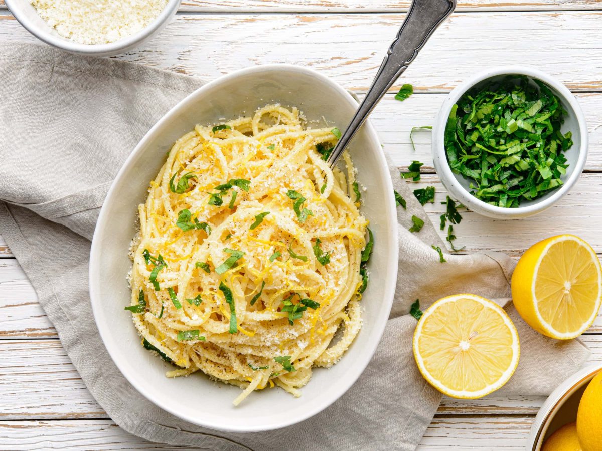 Pasta al Limone in einer weißen Schale. Daneben verschiedene Zutaten. Das Ganze auf einem hölzernen Untergrund.