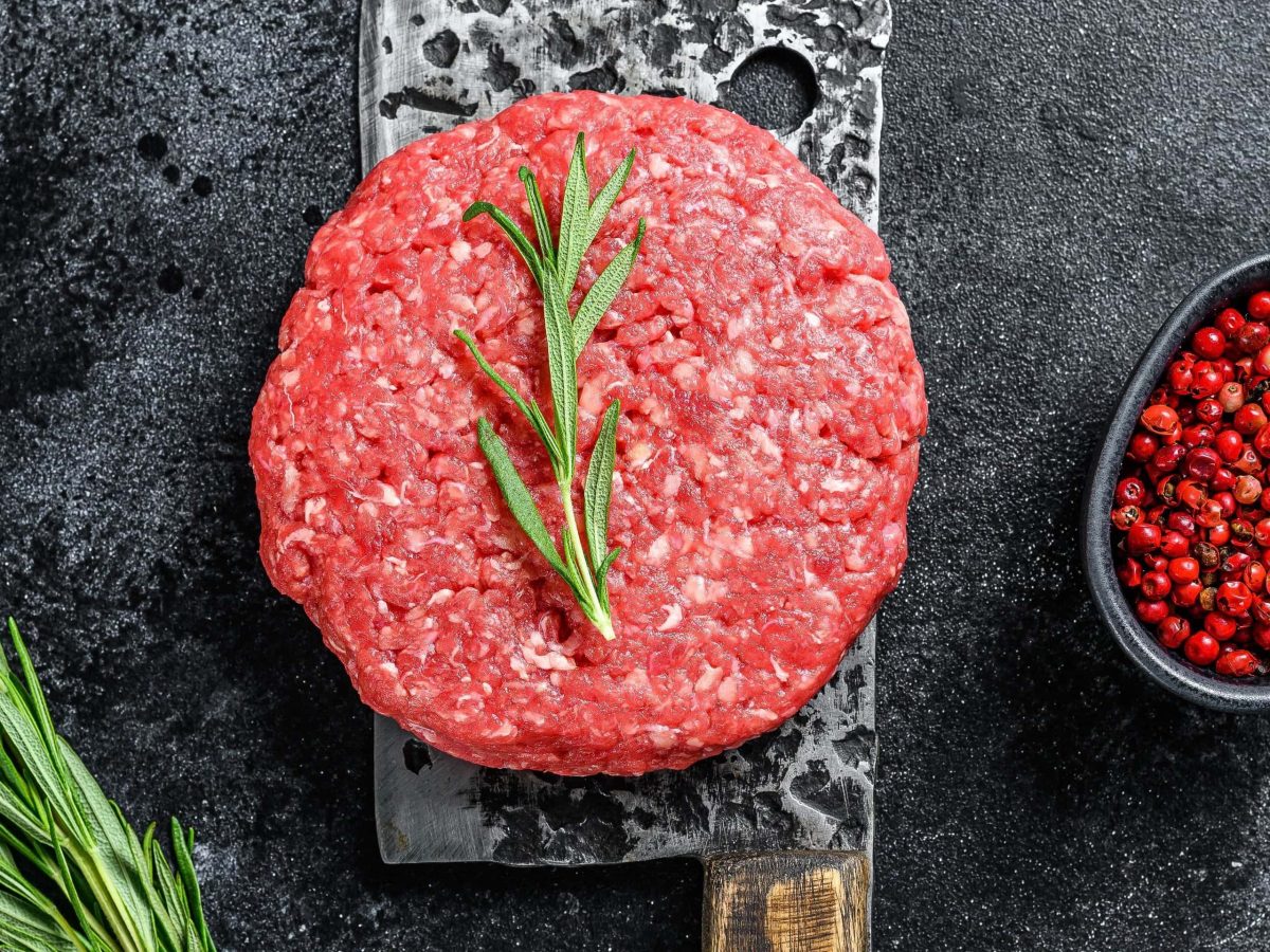 Ein roher Burger-Patty in der Draufsicht auf einem Beil liegen d auf einem grauen Tisch. Rosmarin und roter Pfeffer als Deko-Elemente.