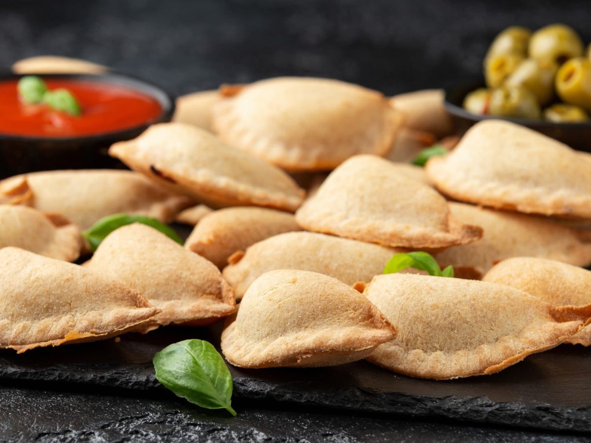 Pizza fritta napoletana auf Schieferplatte. Schwarze Schale mit Tomatensauce und eine weitere mit grünen Oliven im Hintergrund. Basilikumblätter als Dekoration. Frontalansicht.