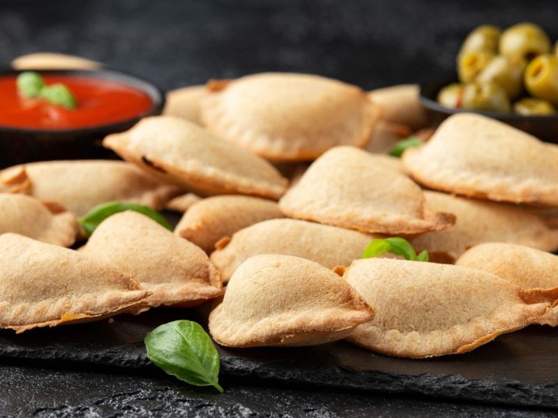 Pizza fritta napoletana auf Schieferplatte. Schwarze Schale mit Tomatensauce und eine weitere mit grünen Oliven im Hintergrund. Basilikumblätter als Dekoration. Frontalansicht.