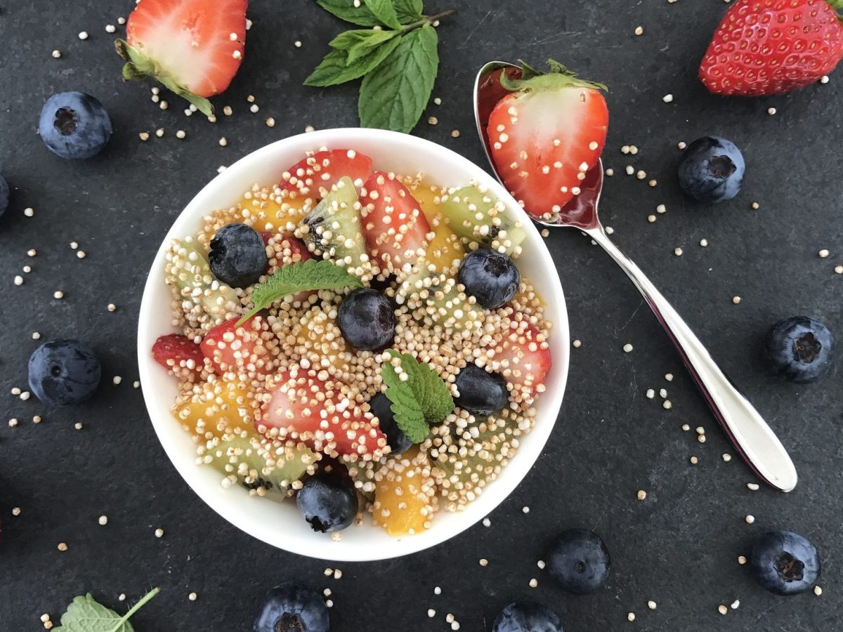 Unser Quinoa-Obstsalat steht angerichtet in einer Schüssel auf einem schwarzen Hintergrund, umringt von Erdbeeren und Blaubeeren.