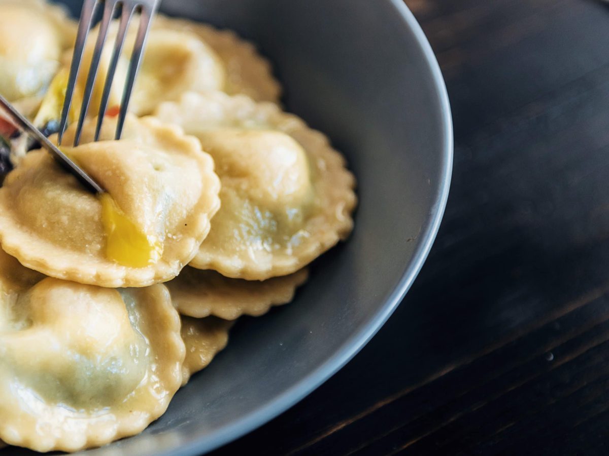 Ravioli in einer grauen Schale und eine Gabel, die in einen hineinsticht. alle spräsentiert auf einem schwarzem Tisch.