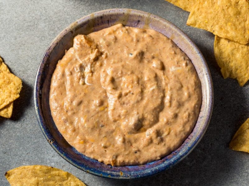 Refried Baked Beans Bohnendip in grauer Schüssel auf grauem Untergrund. Nachos als Dekoration im Bild verteilt. Aufnahme aus der Vogelperspektive.