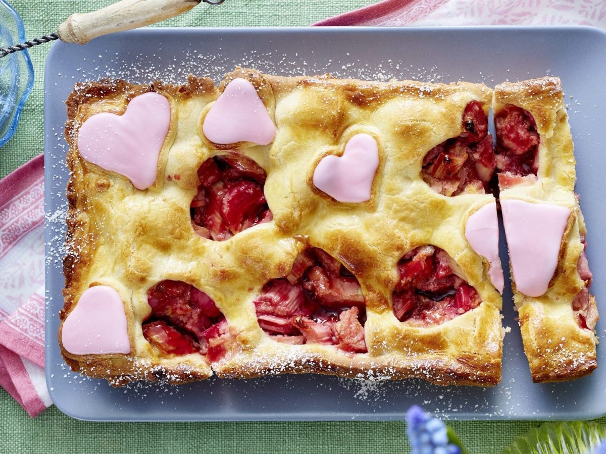 Ein Rhabarber Pie mit Herzen steht mit Puderzucker bestreut auf einer hellen Form. Aus der Teigdecke sind einige Herzen ausgeschnitten. Weitere rosa Herzen sind auf dem Pie verteilt.