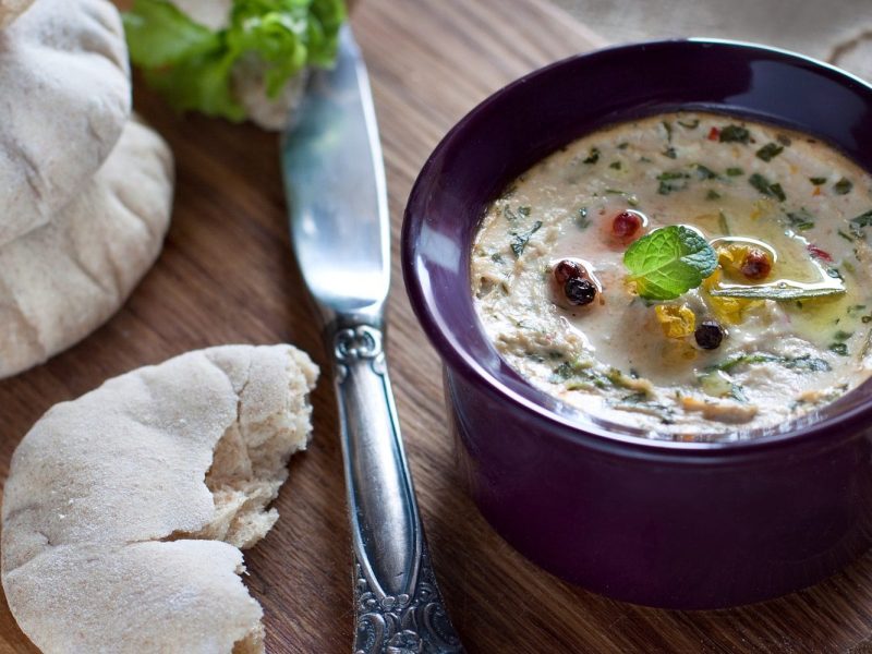 Aus der Vogelperspektive: In eriner kleinen Schlüssel ist ein gebackener Ricotta mit Kräutern. Daneben liegen zwei Flasenbrote, eine Gabel sowie Basilikum-Blätter.