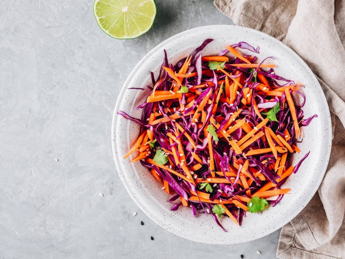 Eine Schüssel Rotkohl-Möhrensalat mit Sesam und Petersilie in der Draufsicht, daneben liegen ein Geschirrtuch und eine halbe Limette.