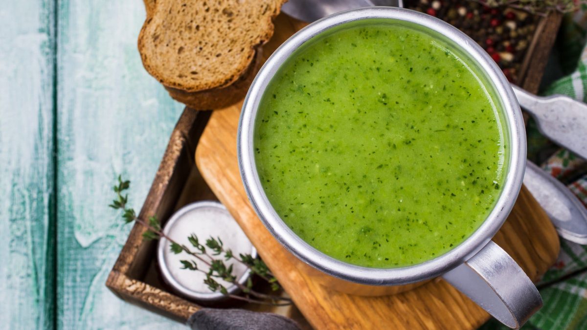 Eine Tasse Rucola-Kartoffel-Suppe in der Draufsicht, sie steht auf einem Holzbrett, daneben liegt eine Scheibe Brot.