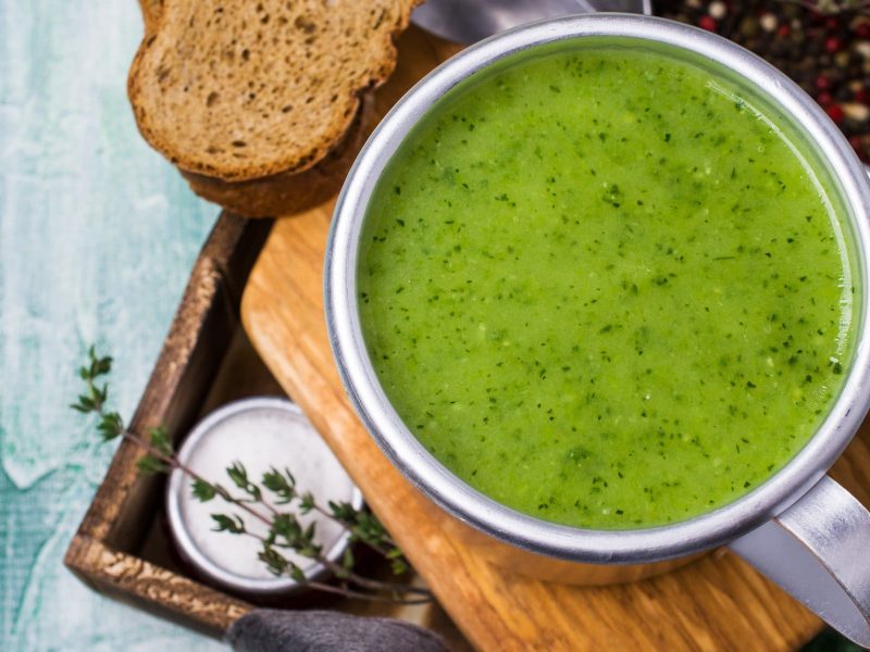 Eine Tasse Rucola-Kartoffel-Suppe in der Draufsicht, sie steht auf einem Holzbrett, daneben liegt eine Scheibe Brot.