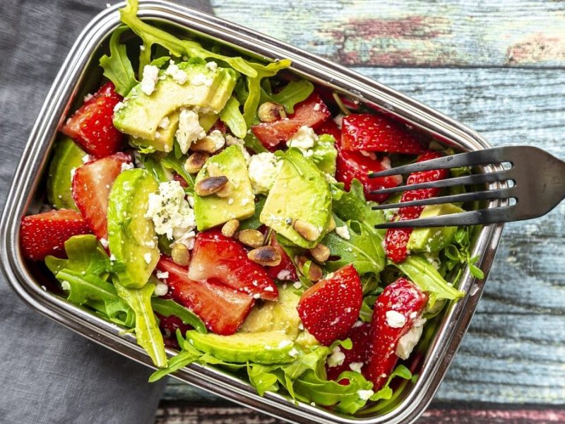 Auf einem bläulichen Untergund steht eine längliche Dose au Metall. Diese hat keinen deckel. In dem Behältnis ist ein Rucola-Salat mit Erdbeeren und Avocado. In dem Salat steckt eine Gabel.