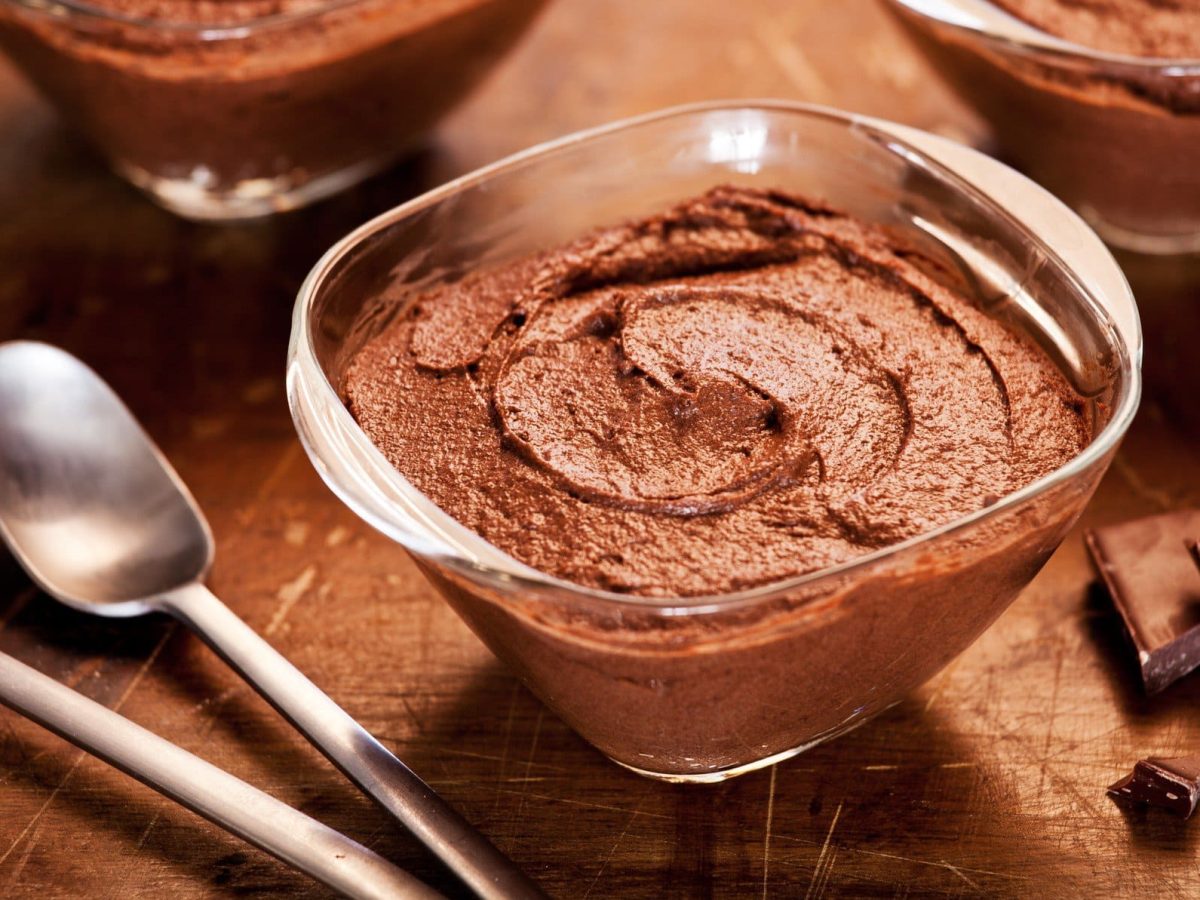 Schnelle Mousse au Chocolat in Glasschale auf braunem Holztisch. Im Hintergrund zwei weitere angedeutet. Links Teelöffel, rechts Schokoladenstücke. Draufsicht.