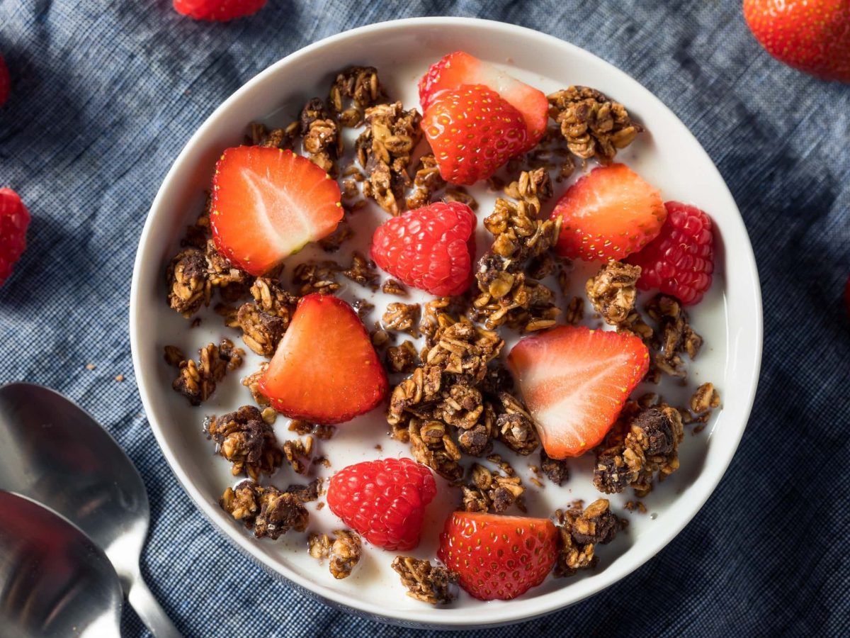 Knuspriges selbst gemachtes Schoko-Granola in einer Schale mit frischen Erdbeeren und Milch, serviert auf einem dunklen Tuch.