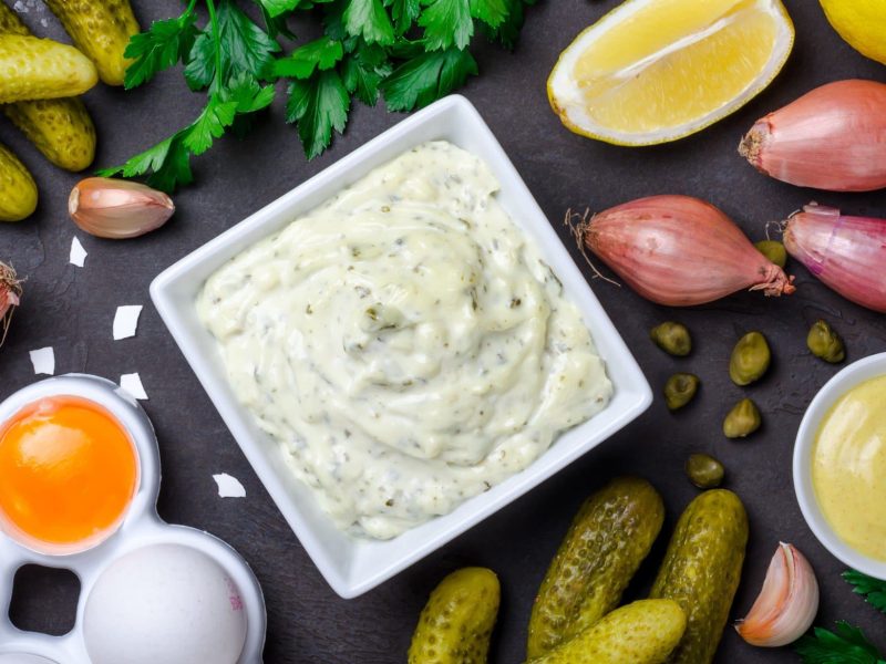 Selbst gemachte Remoulade in einer kleinen Schale. Daneben verschiedene Zutaten auf einem dunklen Untergrund.