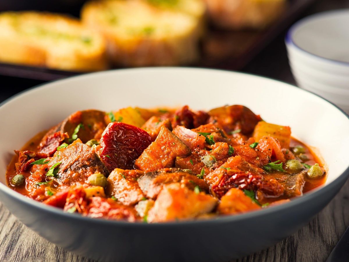 Sizilianische Caponata mit Aubergine, getrockneten Tomaten, Paprika und frischer Petersilie in einer großen Schale auf rustikalem Holztisch.