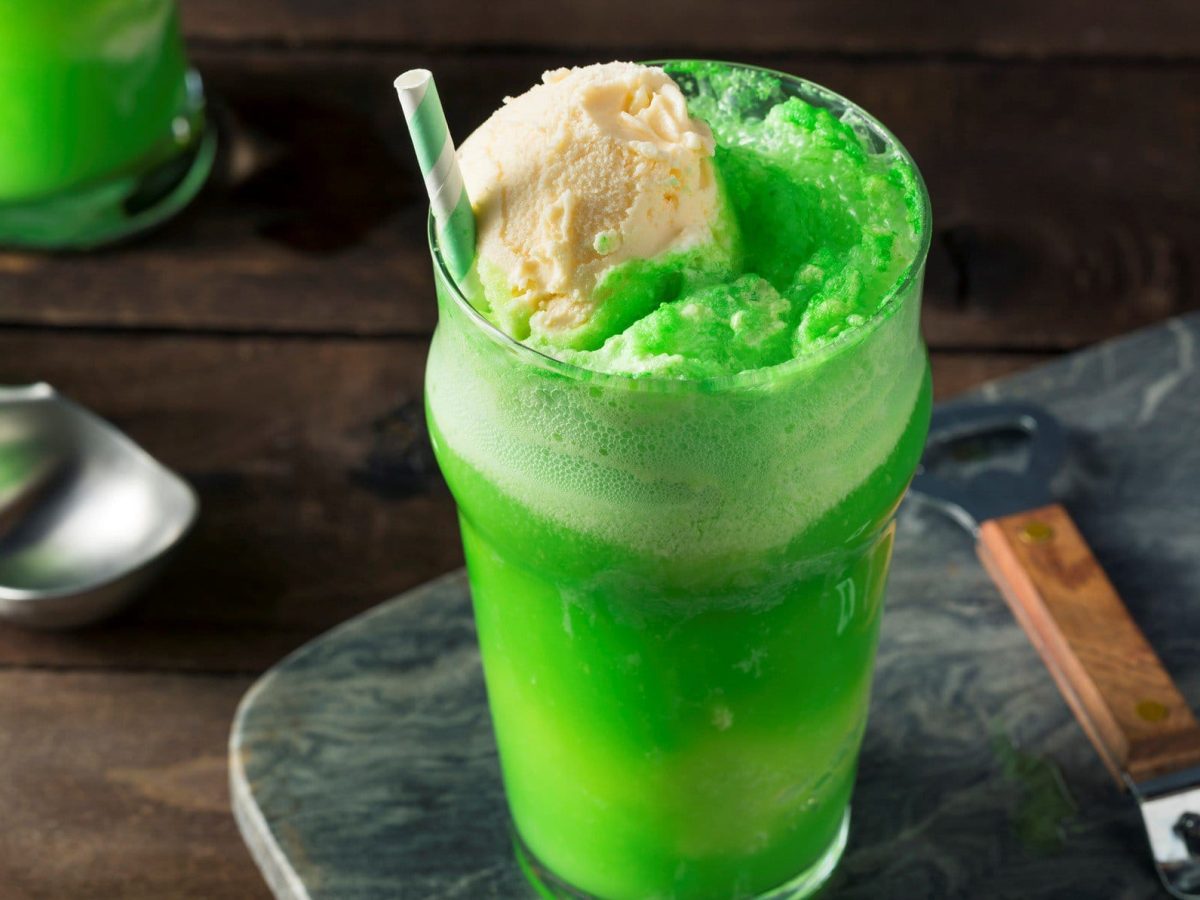 St. Patrick's Day Green Soda Float im Glas mit Strohhalm auf grauer Platte. Daneben Löffel mit Holzgriff. Links Eislöffel und ein weiteres Glas angedeutet. Frontalaufnahme.