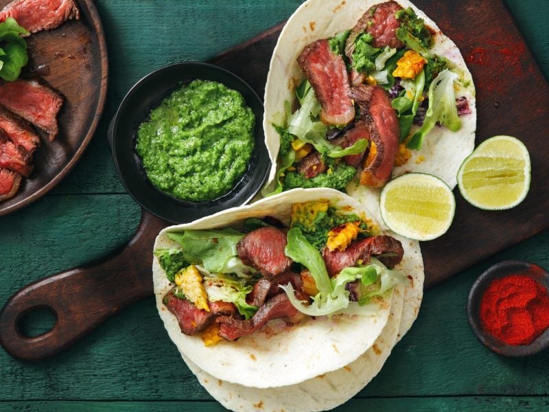 Steak Tacos mit grünem Pesto auf einem Holzbrett. Daneben verschiedene Zutaten auf einem blauen hölzernen Untergrund.