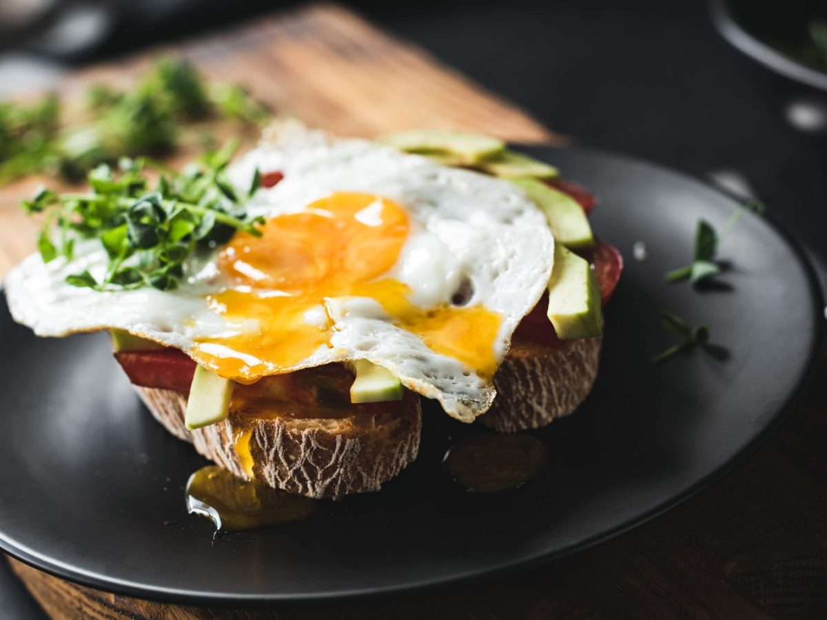 Schwarzer Teller in der Draufsicht mit Brot, Avocado, Sucuk und einem Spiegelei