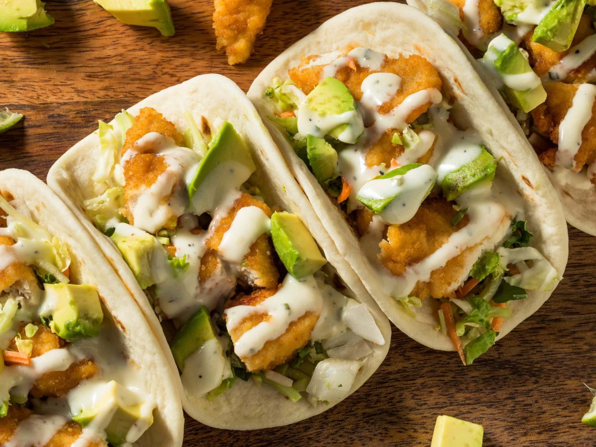 Fried Chicken Tacos von oben mit paniertem Hähnchen und Avocado Stücken auf Holztisch.