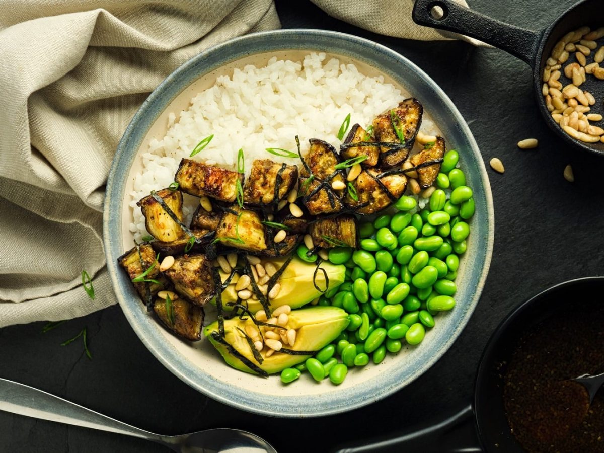 Teriyaki-Bowl mit Aubergine, Edamame, Avocado, Reis und Pinienkernen auf dunklem Untergrund in der Draufsicht, daneben ein Geschirrtuch und eine Schale Pinienkerne.