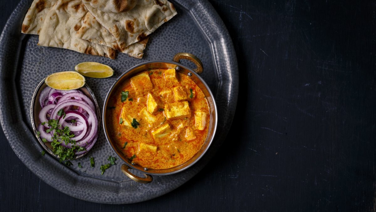 Ein Tablett mit einer Schüssel Tofu nach Butter-Chicken-Art, Naanbrot und Zwiebel in der Draufsicht.