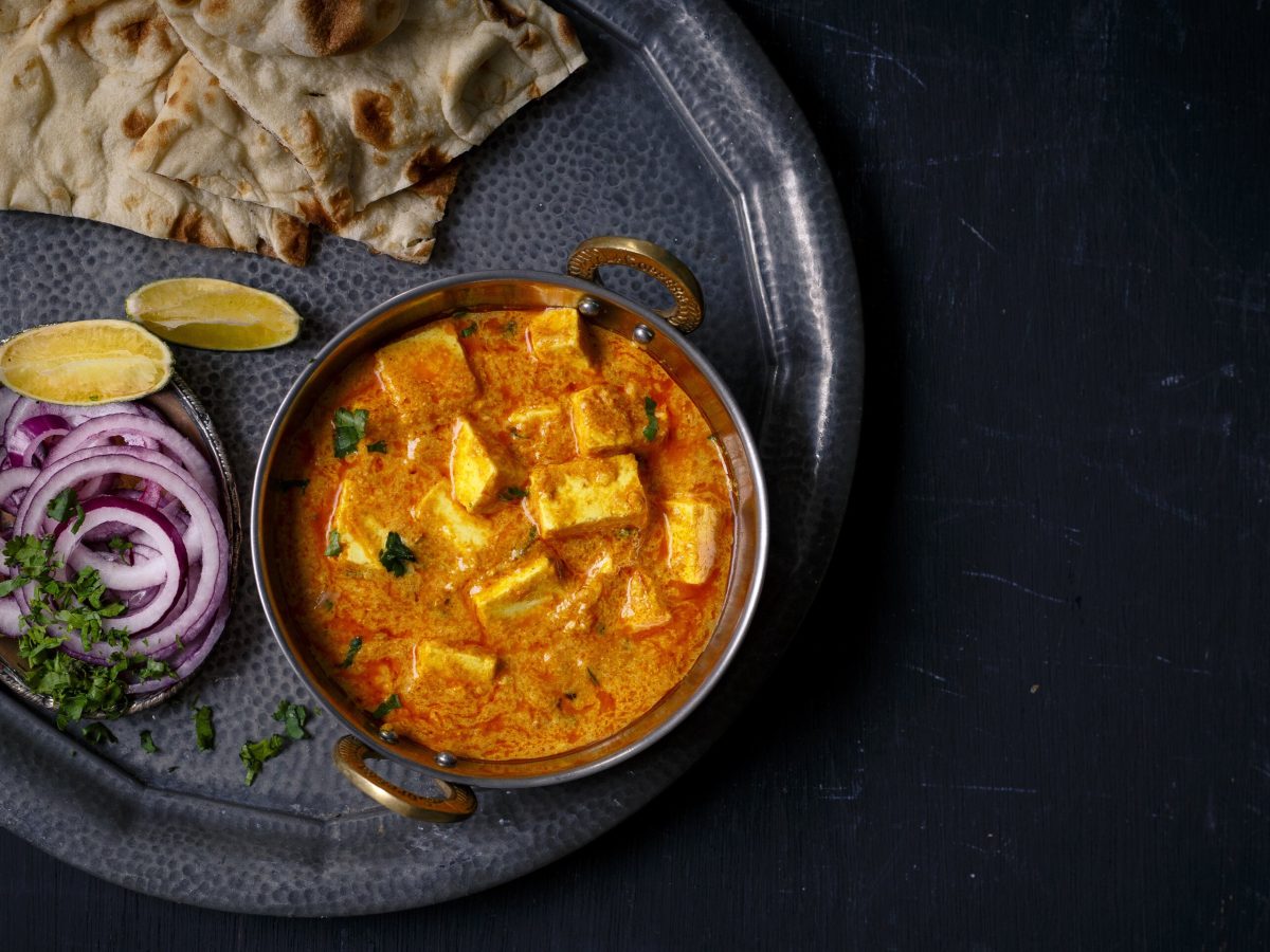 Ein Tablett mit einer Schüssel Tofu nach Butter-Chicken-Art, Naanbrot und Zwiebel in der Draufsicht.