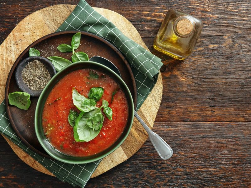 Eine Schüssel Tomaten-Sellerie-Suppe mit Basilikum auf Holz in der Draufsicht, daneben steht eine Flasche Öl.