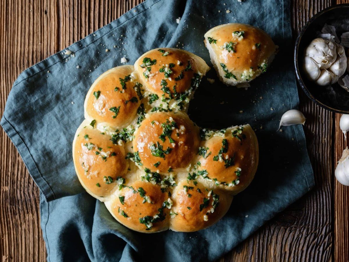 Ukrainische Pampushka auf einem blauen Küchentuch. Daneben eine Schale mit Knoblauch. Das Ganze auf einem hölzernen Untergrund.