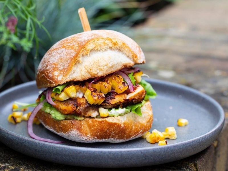 Ein veganer Elote-Burger mit Mais, Salat und selbst gemachtem Bratling auf einem grauen Teller.