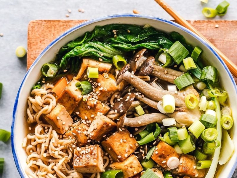 Eine Schale vegane Miso-Ramen mit Räuchertofu, Frühlingszwiebeln, Pilzen, Nudeln, Pak Choi und Sesam in der Draufsicht, daneben liegen Essstäbchen.