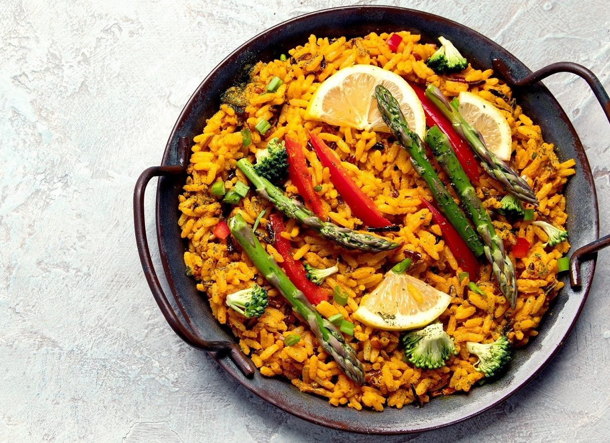 Eine gusseiserne Pfanne mit veganer Paella mit grünem Spargel, Paprika, Brokkoli und Zitronenscheiben.