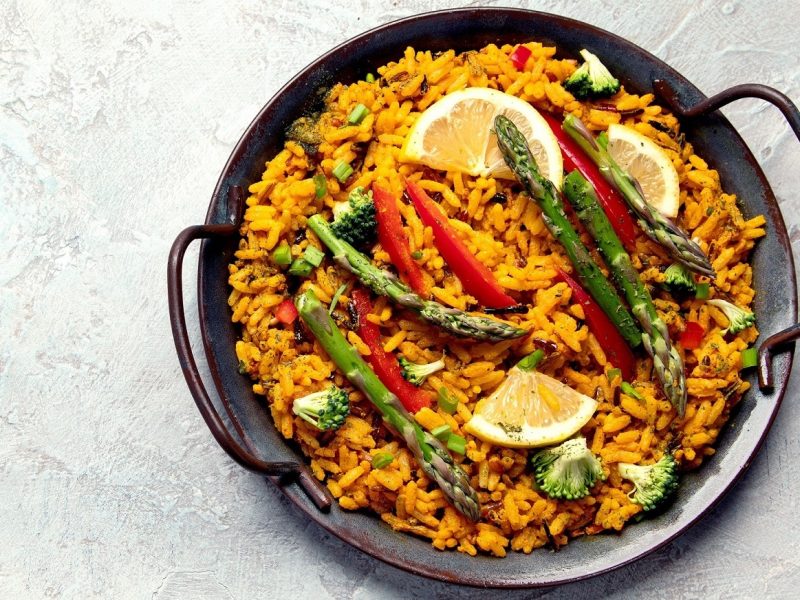 Eine gusseiserne Pfanne mit veganer Paella mit grünem Spargel, Paprika, Brokkoli und Zitronenscheiben.