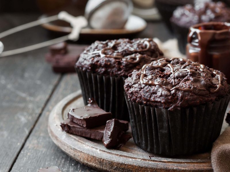 Zwei vegane Schoko-Himbeer-Muffins auf einem runden Holzbrett, daneben liegen Kuvertüre-Stücke, im Hintergrund sind unscharf Küchenutensilien zu sehen.