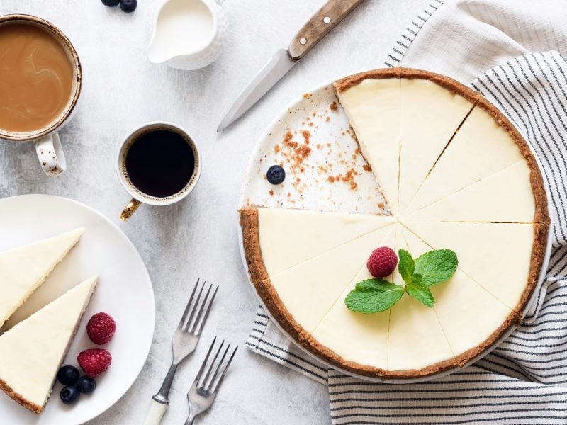 Ein angeschnittener veganer New York Cheesecake in der Draufsicht, daneben steht ein Teller mit zwei Stück Kuchen sowie zwei Kaffeetassen und ein Milchkännchen.