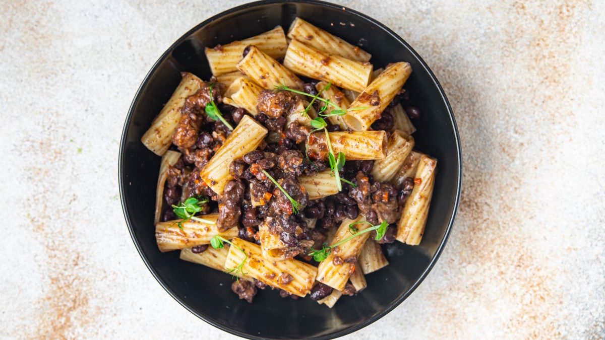 Ein schwarzer Teller veganes Linsenragout mit Rigatoni und frischen Kräutern auf hellem Marmor in der Draufsicht.