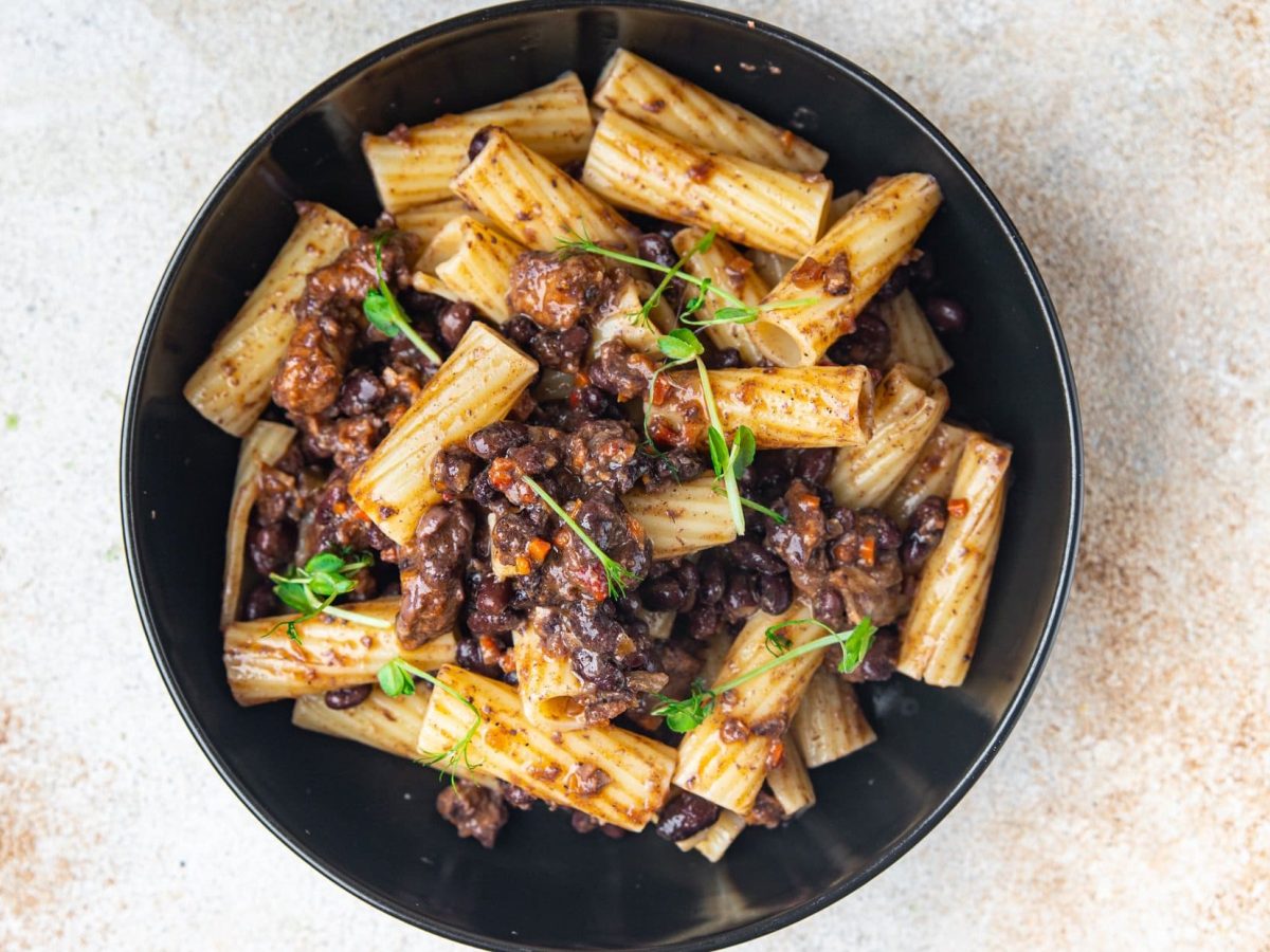 Ein schwarzer Teller veganes Linsenragout mit Rigatoni und frischen Kräutern auf hellem Marmor in der Draufsicht.