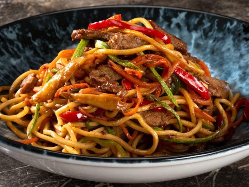 Wafu Miso Steak mit Gemüse und Udon-Nudeln in schwarz-weißer Schale auf Holztisch. Frontalaufnahme.