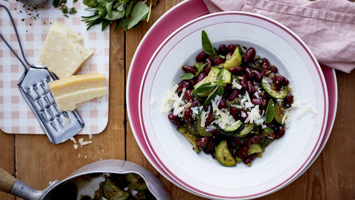 Unser Zucchini-Bohnen-Topf steht fein angerichtet in einem flachen Teller auf dem Tisch. Daneben liegen Parmesan und Kräuter.