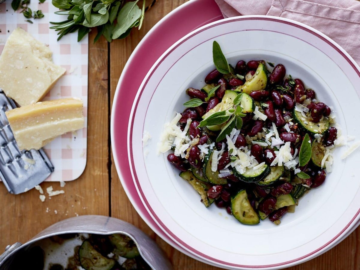 Unser Zucchini-Bohnen-Topf steht fein angerichtet in einem flachen Teller auf dem Tisch. Daneben liegen Parmesan und Kräuter.