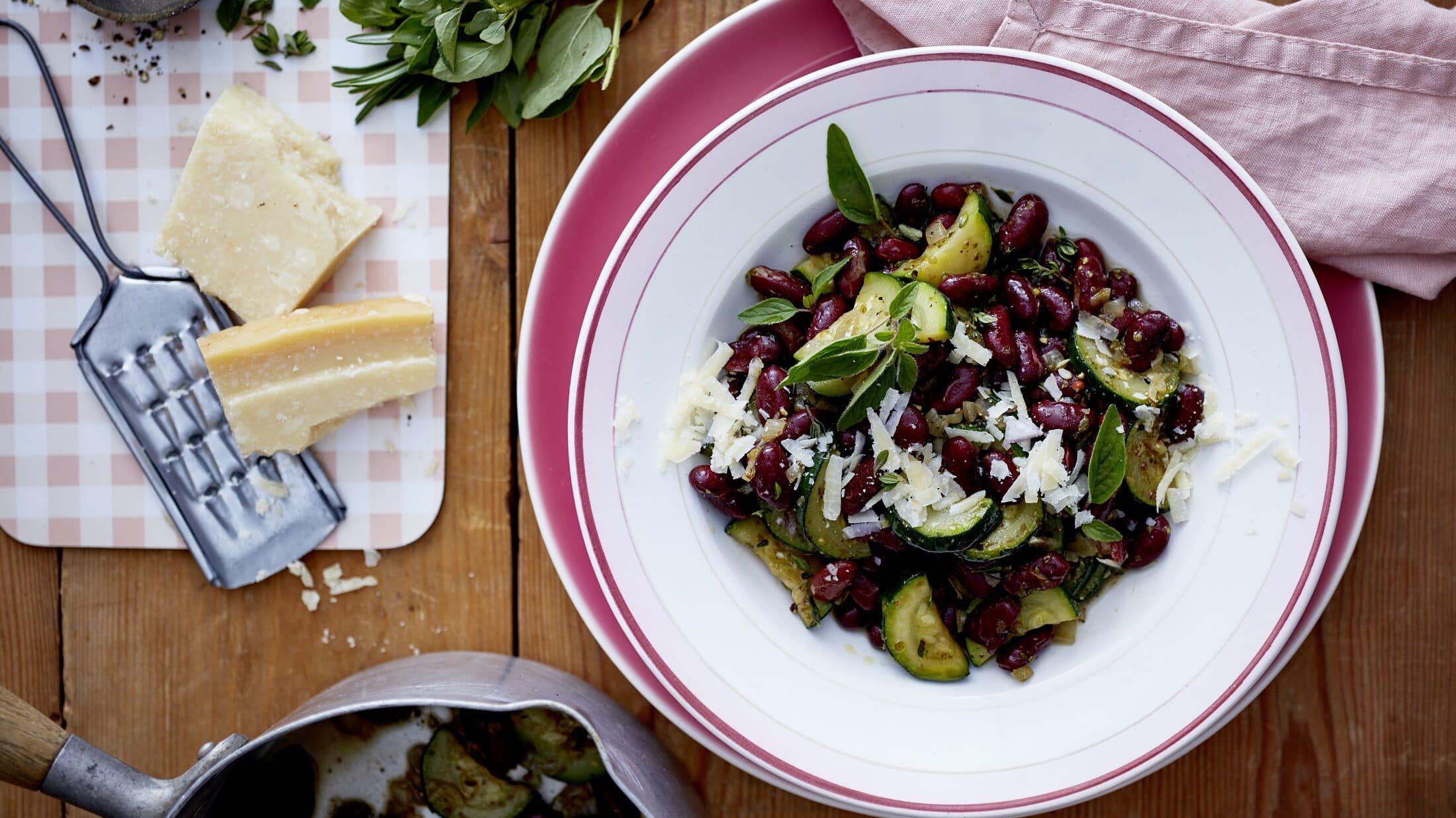 Unser Zucchini-Bohnen-Topf steht fein angerichtet in einem flachen Teller auf dem Tisch. Daneben liegen Parmesan und Kräuter.