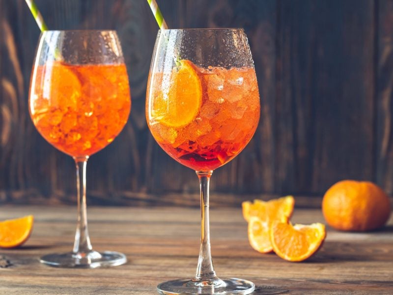 Zwei Gläser mit Aperol Spritz auf hellem Holzuntegrund. Dabeben liegen angeschnittene Orangen.