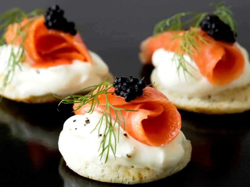 Blini mit Lachs, Sauerrahm und Kaviar dekoriert mit frischem Dill auf glänzendem, dunklen Untergrund.