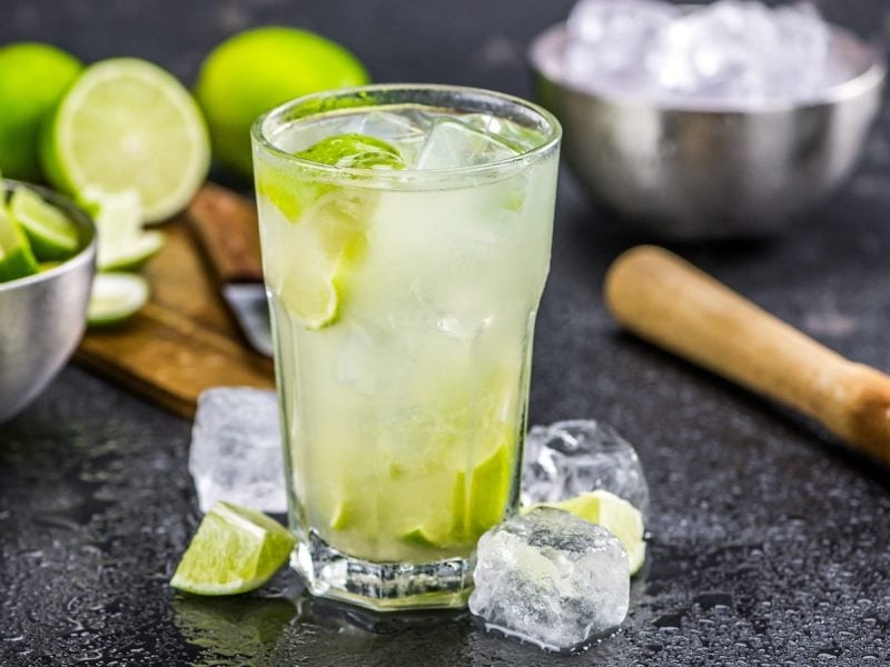 Ein Glas Caipirinha auf dunklem Untergrund. Daneben steht eine Schale mit Limettenwürfeln und eine mit Eiswürfeln. Außerdem ein Brett mit Limetten sowie ein Stößel