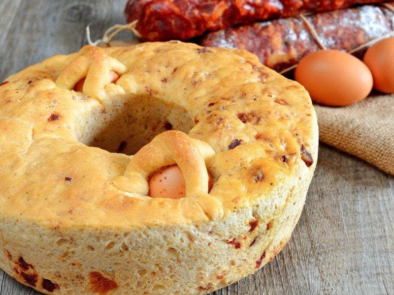 Ein mit hart gekochten Eiern gefüllter, herzhafter Osterkranz aus Italien auf hölzernem Untergrund. Daneben liegen Zutaten wie Eier und Salami.