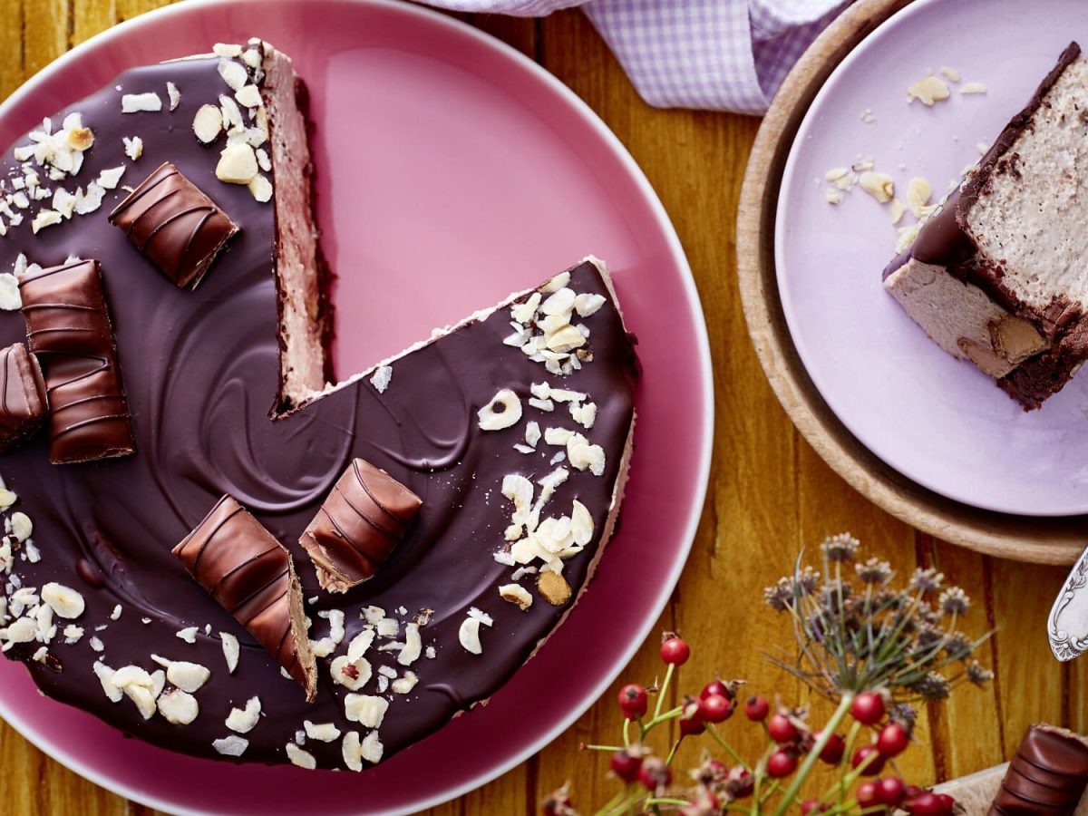 Auf einem rosa Teller liegt ein angeschnittener Haselnuss-Cheesecake. Man erkennt die Creme Schicht in der Mitte. Er ist mit einer Schokoladenglasur überzogen und mit Haselnüssen bestreut.