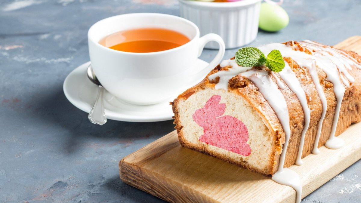 Auf einem Brett liegt ein Osterhasenkuchen aus der Kastenform. Er ist angeschnitten und man erkennt den eingebackenen rosa Hasen im Teig. Der Kuchen ist mit Zuckerguss überzogen.