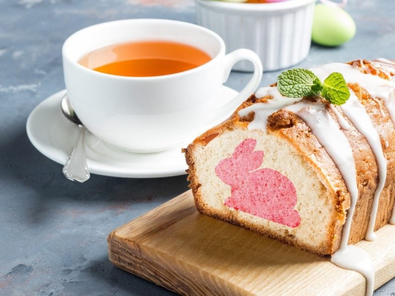 Auf einem Brett liegt ein Osterhasenkuchen aus der Kastenform. Er ist angeschnitten und man erkennt den eingebackenen rosa Hasen im Teig. Der Kuchen ist mit Zuckerguss überzogen.