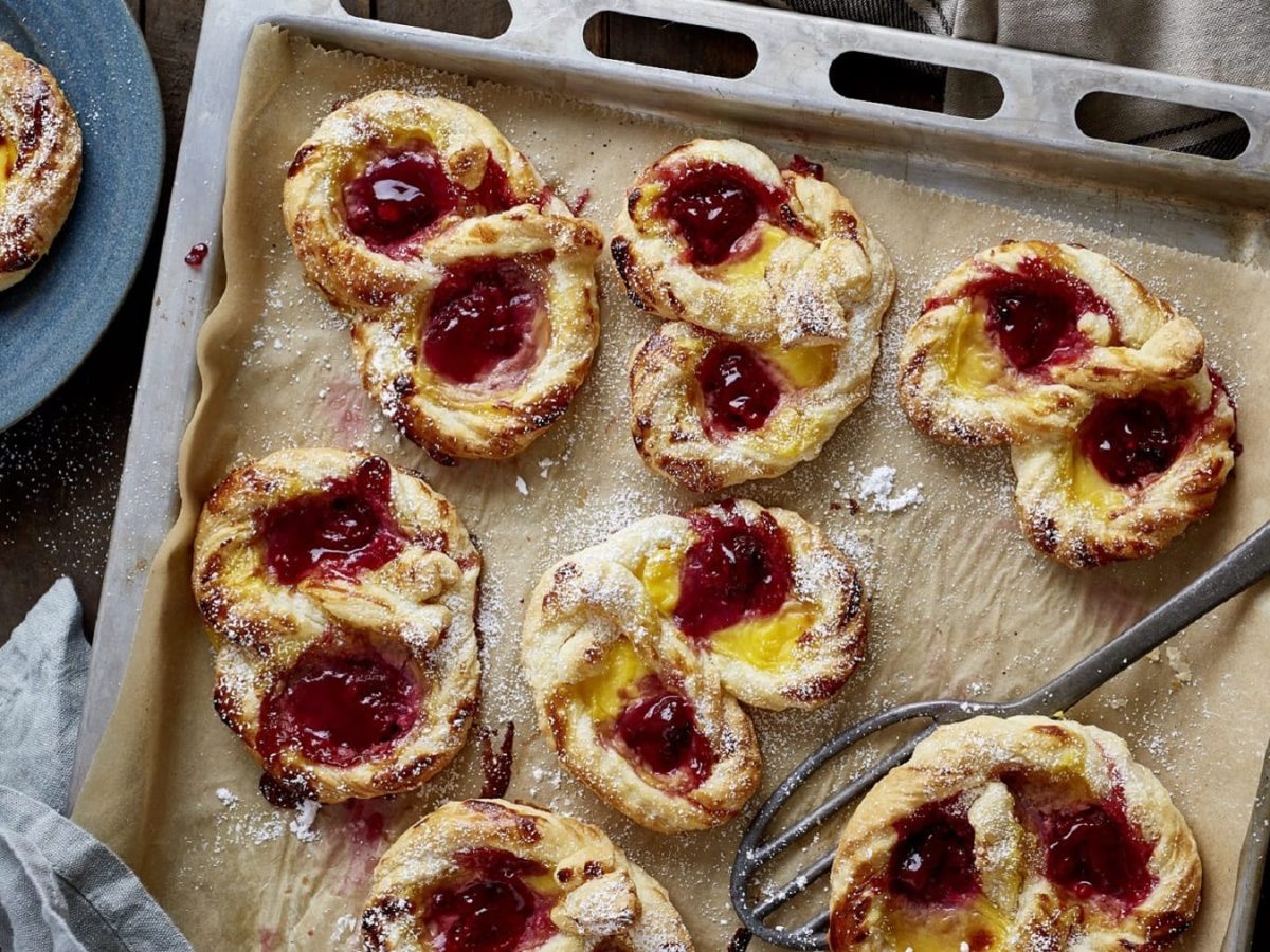 Auf einem Backblech liegen mehrere Pudding-Himbeer-Brezeln. Sie sind aus Blätterteig gemacht und mit Vanillepudding und Himbeerkonfitüre gefüllt.