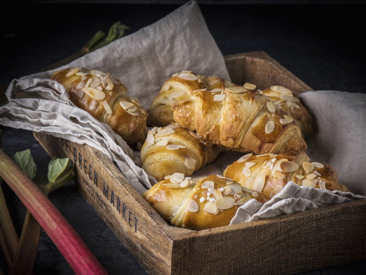 In einem Brotkorb liegen mehrere Rhabarber-Croissants. Sie sind mit Mandelblättchen bestreut und sind goldbraun gebacken.