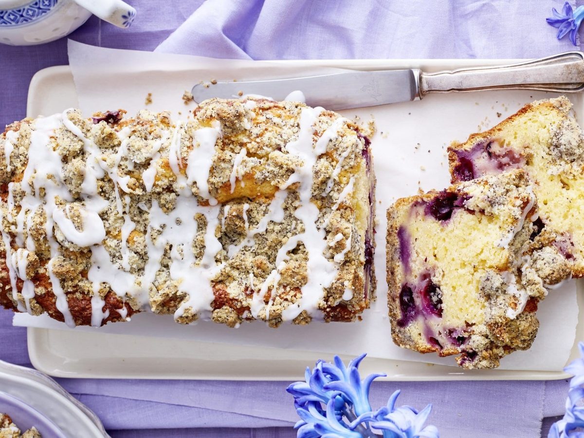 Auf einer Porzellanplatte liegt ein Mohn-Zitronenkuchen aus der Kastenform. Er hat Heidelbeeren im Teig und ist mit Zuckerguss überzogen.