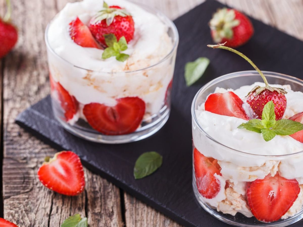 Zwei Gläser mit Eton Mess auf Schieferplatte auf Holzuntergrund. Darum herum liegen Erdbeeren.