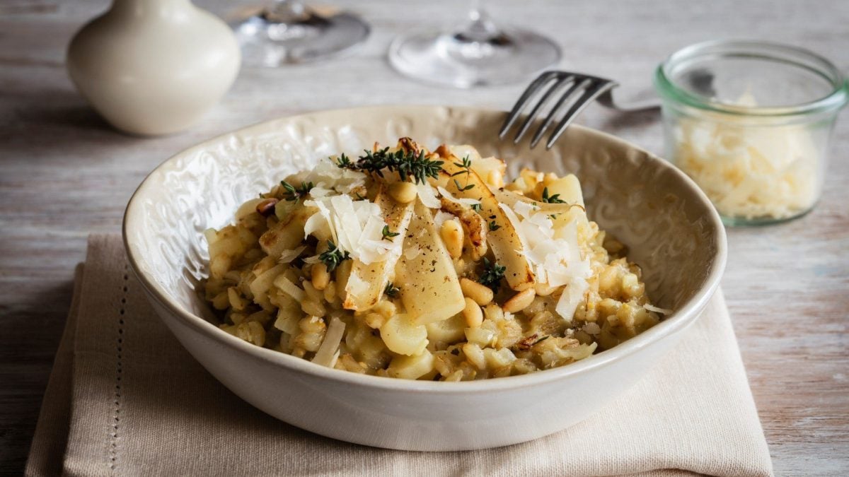 Ein heller Teller mit Gerstenrisotto und Spargelstangen auf einem beigen Untergrund. Dazu ein Glas mit Parmesan und helles Geschirr im Hintergrund.